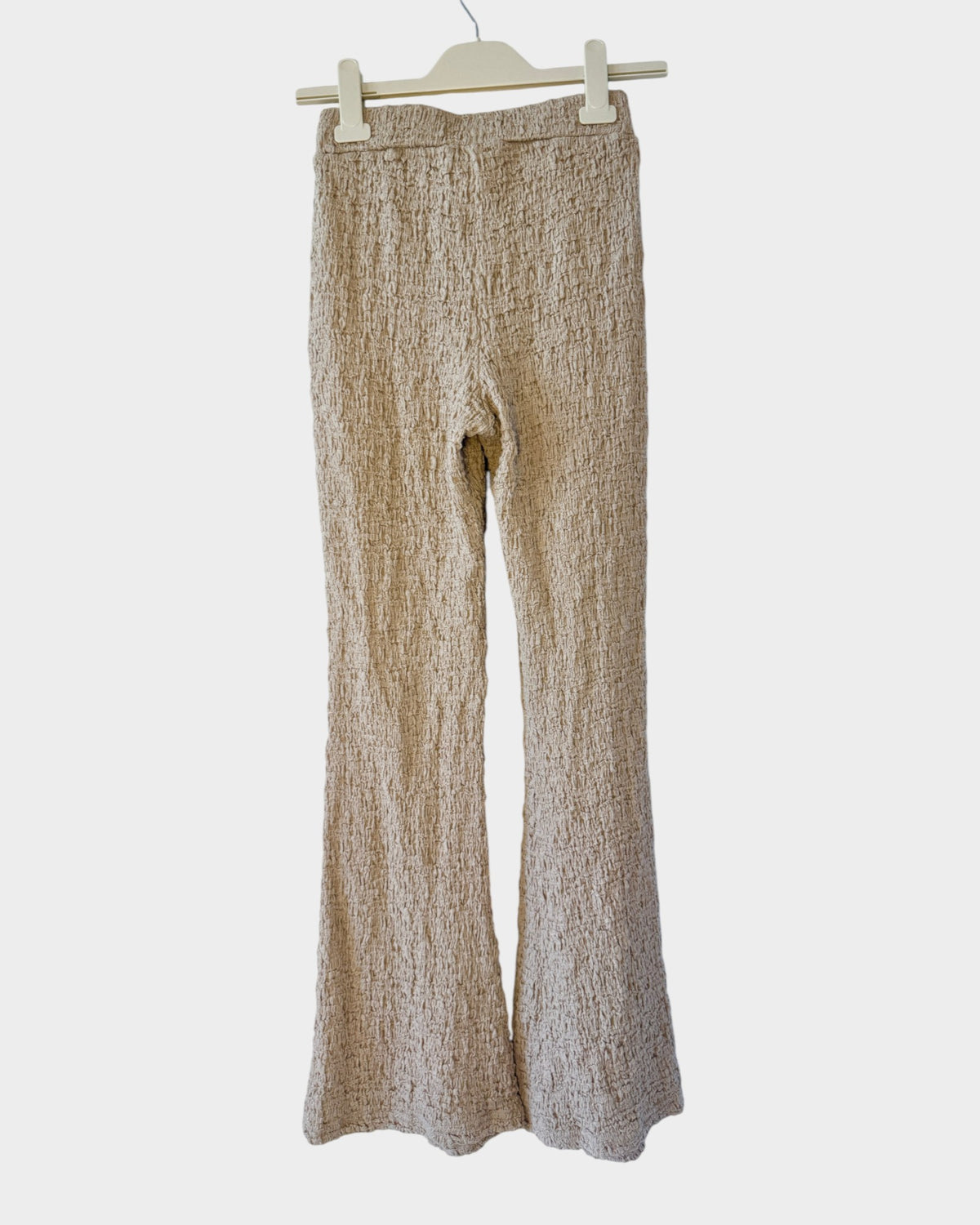 Lula and Mimi beige textured knit trousers with a flared leg design hanging on a white brick wall background.