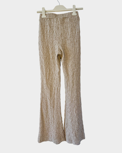 Lula and Mimi beige textured knit trousers with a flared leg design hanging on a white brick wall background.